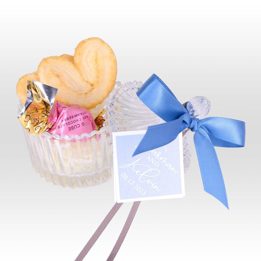 A SWEET MOMENT WEDDING FAVOURS WITH CRYSTAL GLASSWARE BOX- HANDMADE PALMIE & CHOCOLATE cupcake with a blue ribbon and a heart shaped cookie.